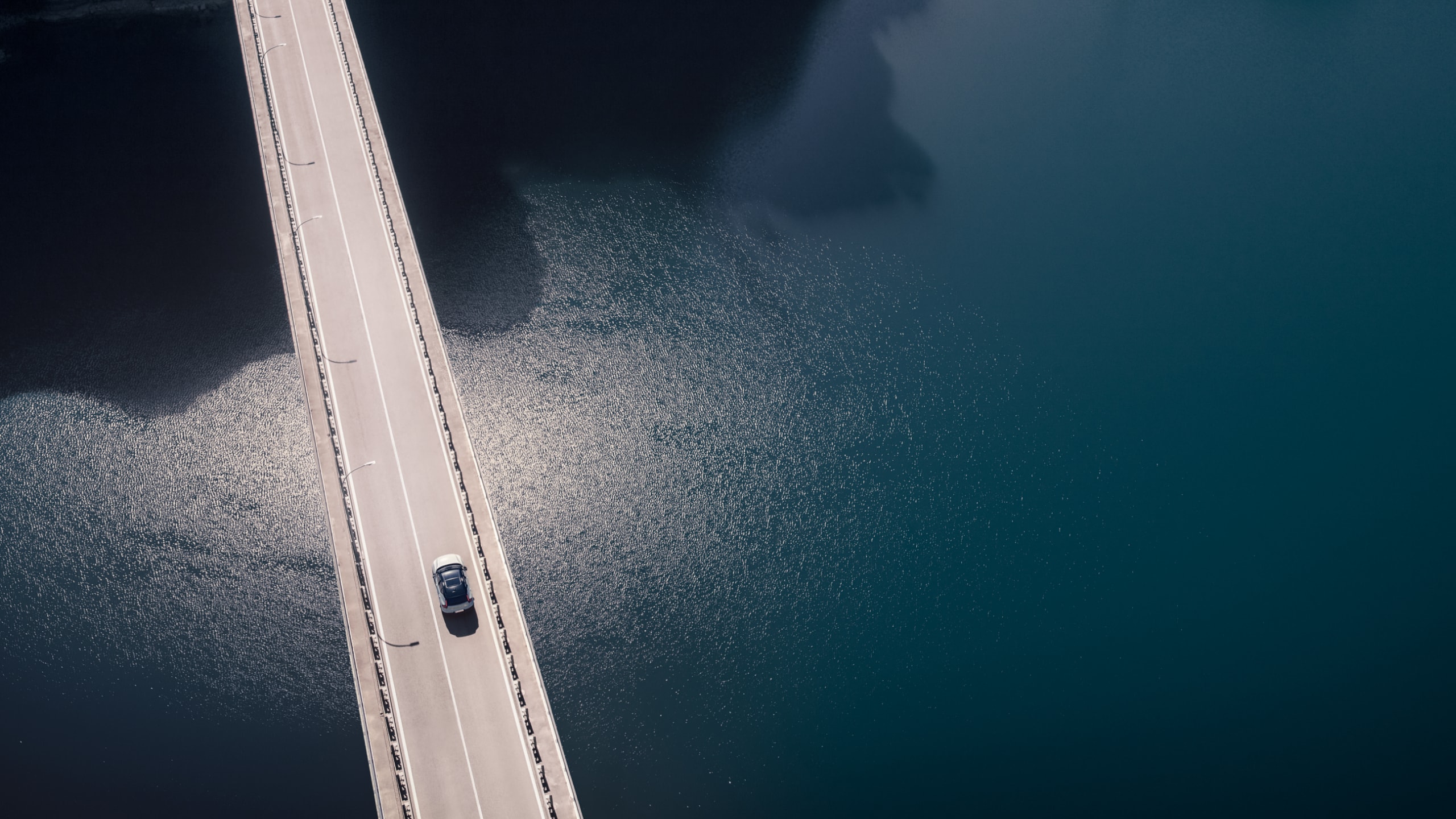 Vista aerea di una nuova Volvo XC40 Recharge Pure Electric SUV che attraversa un ponte a due corsie su acque calme e grigie e si dirige verso un tunnel.