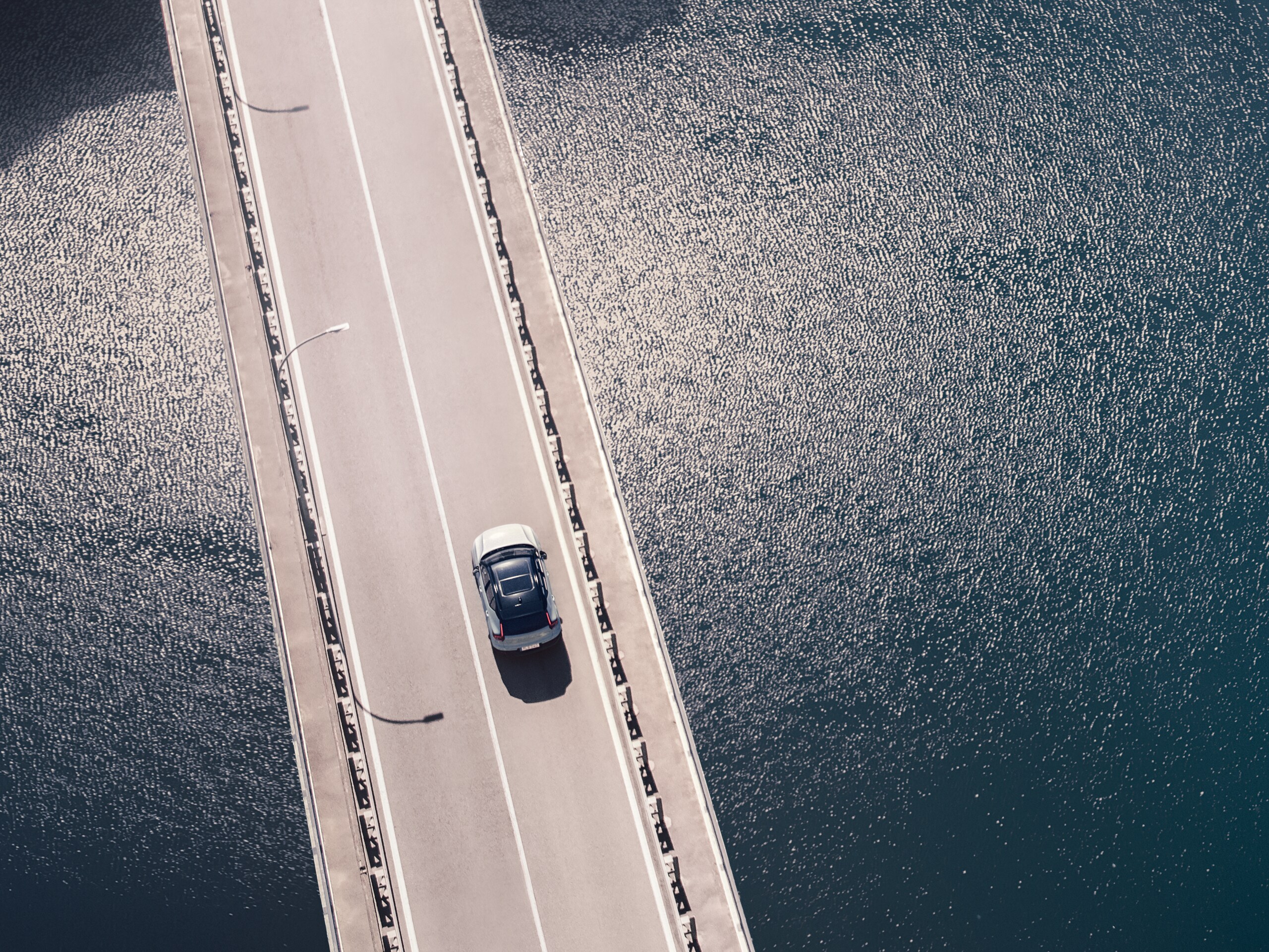 Luftaufnahme eines neuen Volvo XC40 Recharge Pure Electric SUV, das eine zweispurige Brücke über ruhigem, dunkelgrauen Wasser überquert und auf einen Tunnel zufährt.