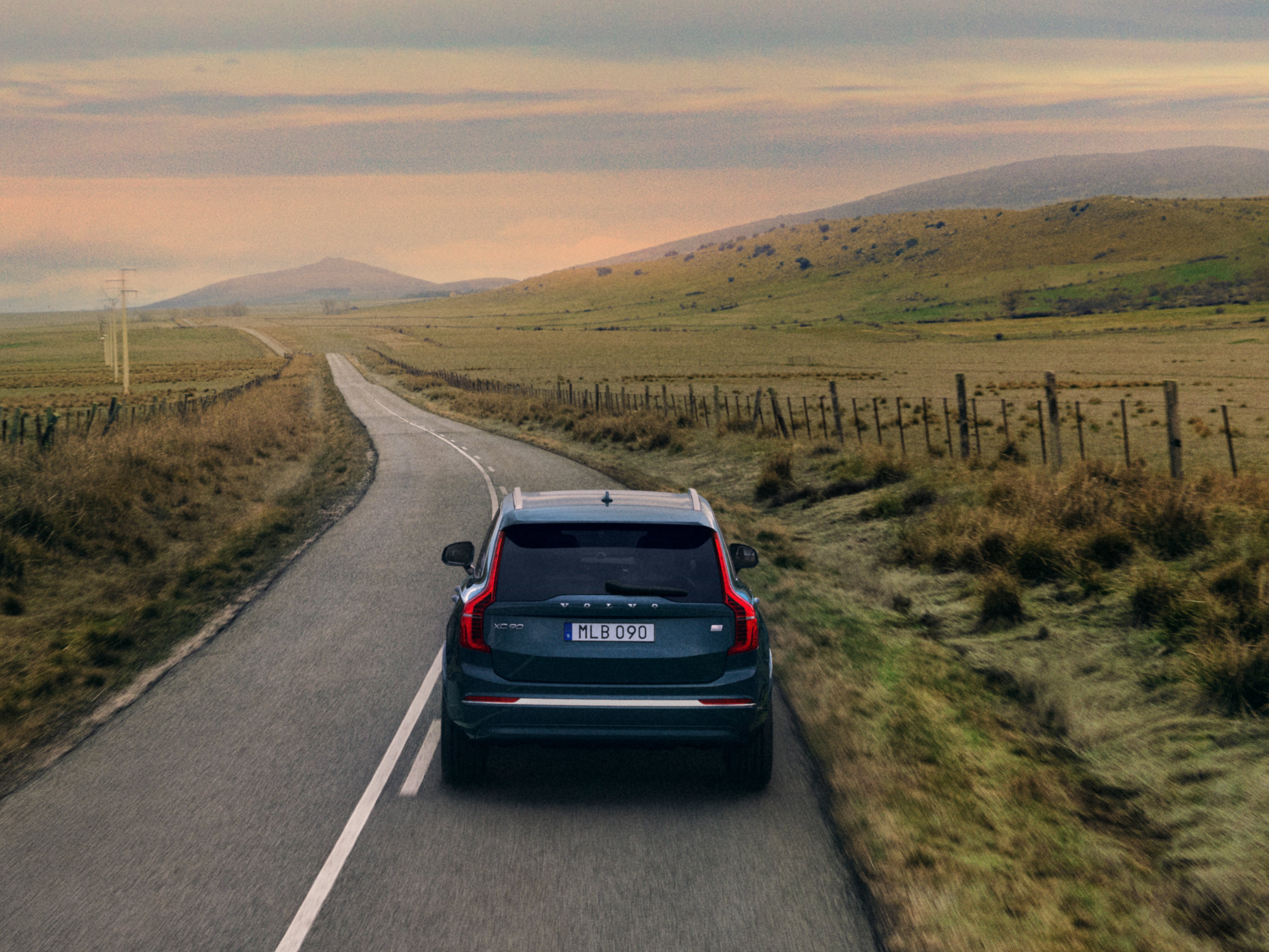 Un Volvo XC90 Recharge híbrido enchufable visto desde atrás circulando por una carretera en el campo.