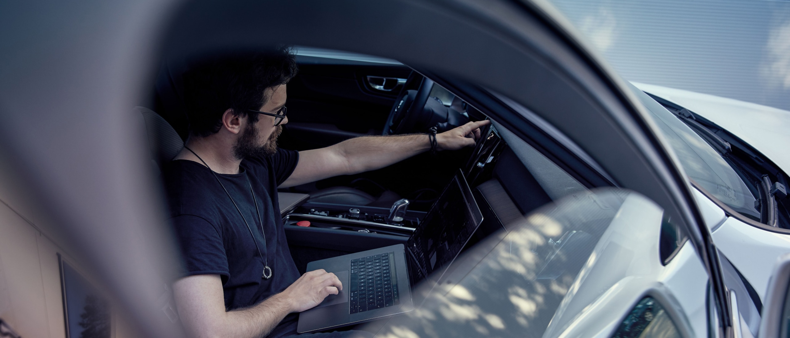 A man sat inside a car working on a laptop with a finger on the in-car system.