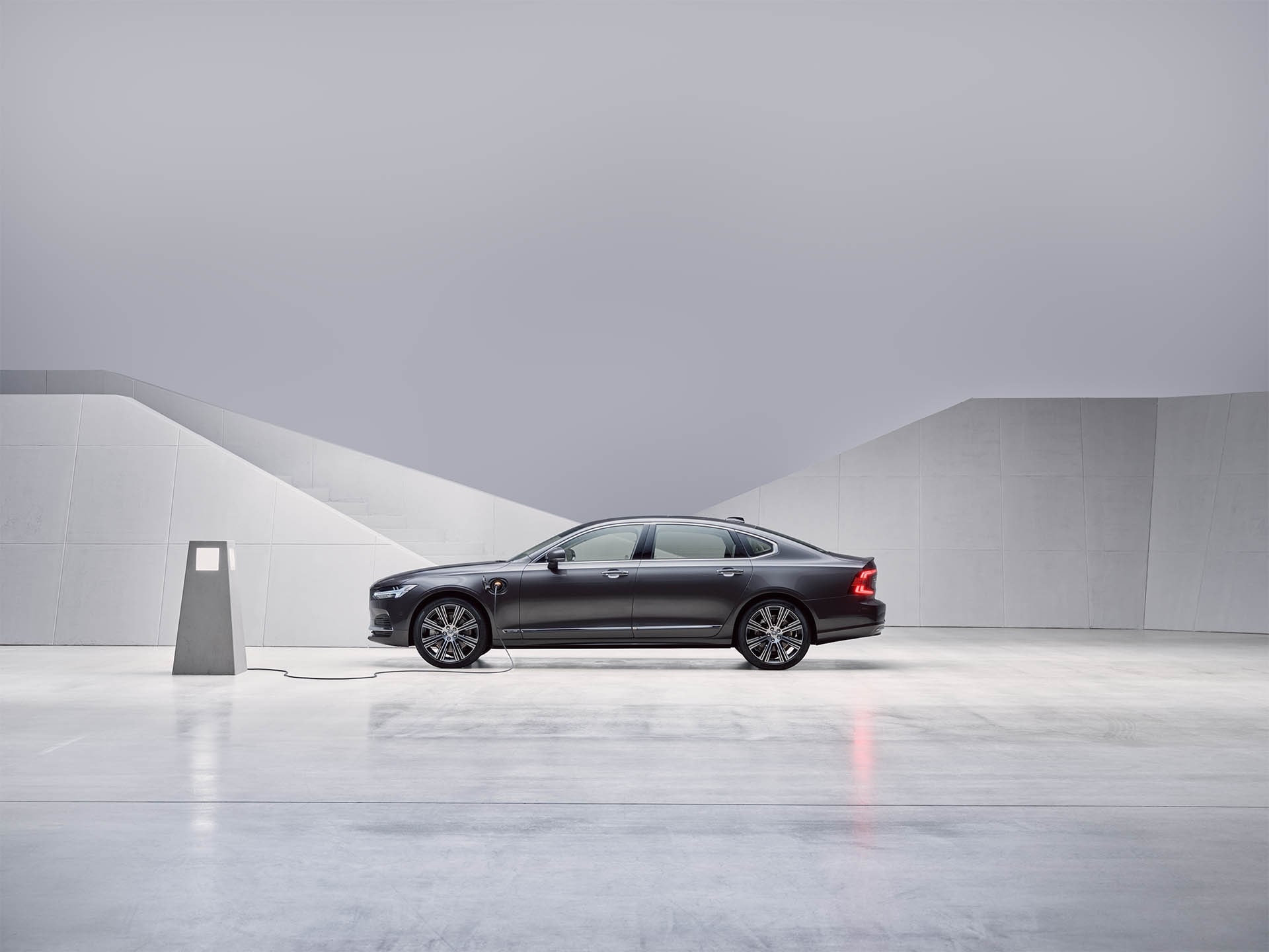 Stojící plug-in hybridní sedan Volvo S90 v šedém odstínu Pebble Grey při nabíjení