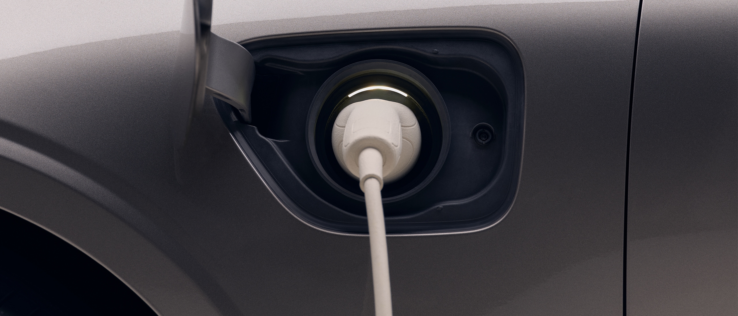 Krupni plan prednje lijeve četvrtine novog tamnosivog Volvo električnog automobila s bijelim kablom za punjenje priključenim u priključak za punjenje automobila.
