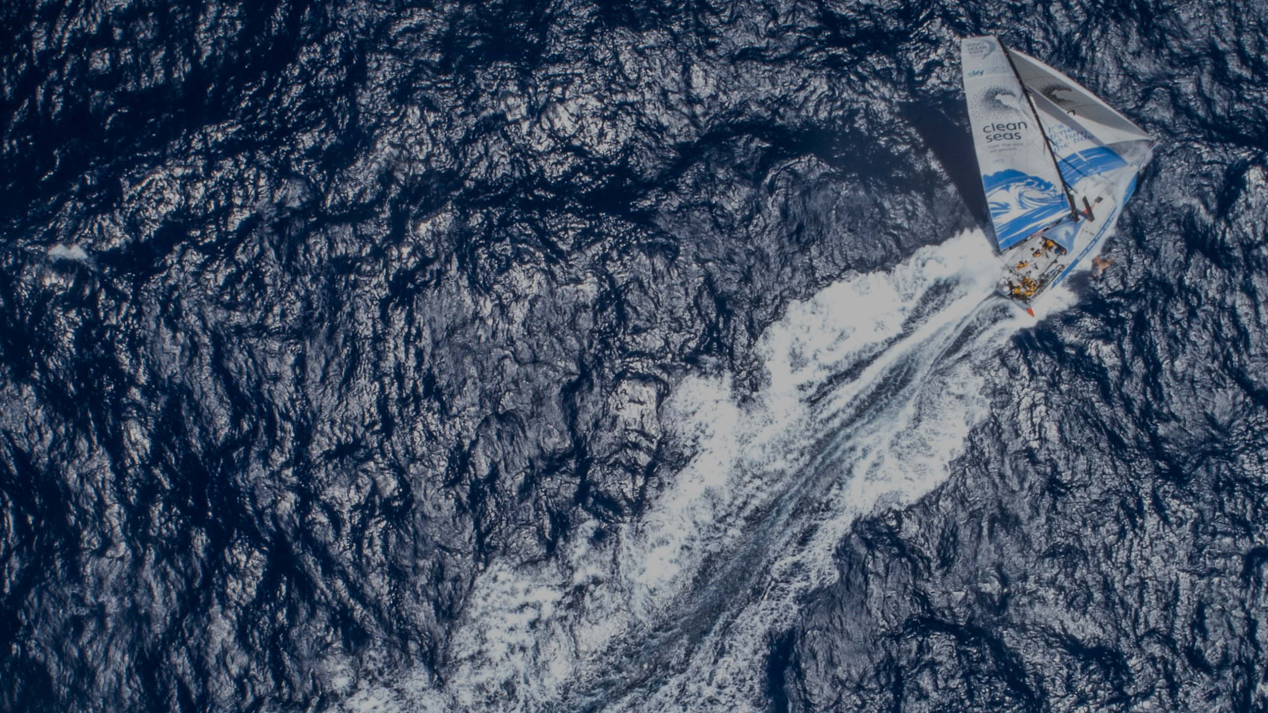 Visão de um pássaro das calmas águas no oceano, com um veleiro no centro deixando um rastro branco ondulado na água azul.
