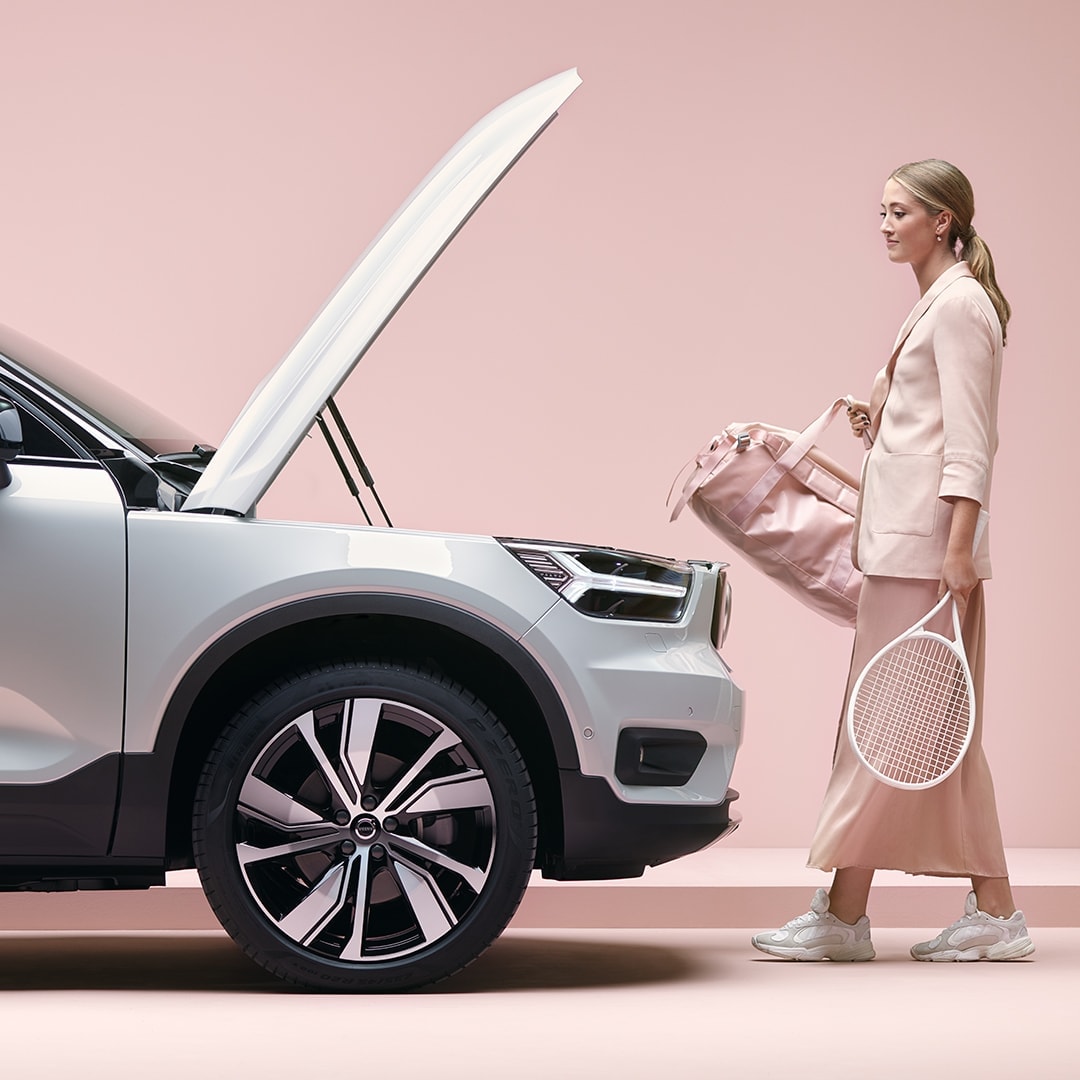 Jeune fille blonde avec un sac de sport et une raquette de tennis à la main en route vers son SUV Volvo XC40 électrique Recharge
