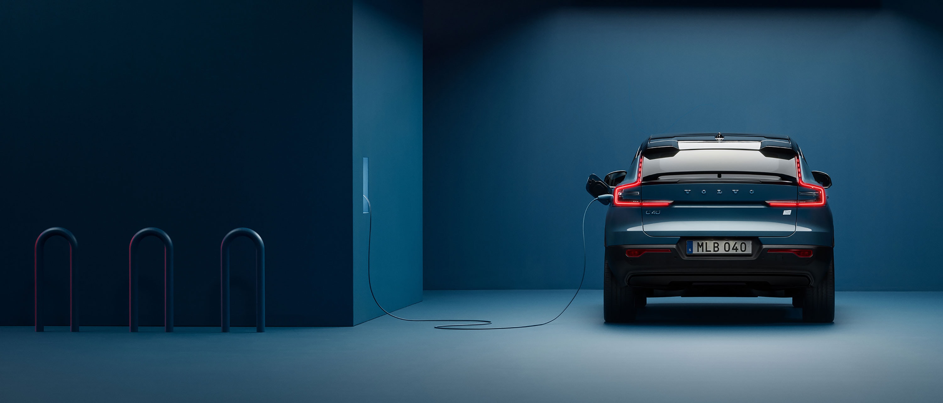 A Volvo C40 Recharge is seen from the rear being charged from a wallbox in a dark blue room.