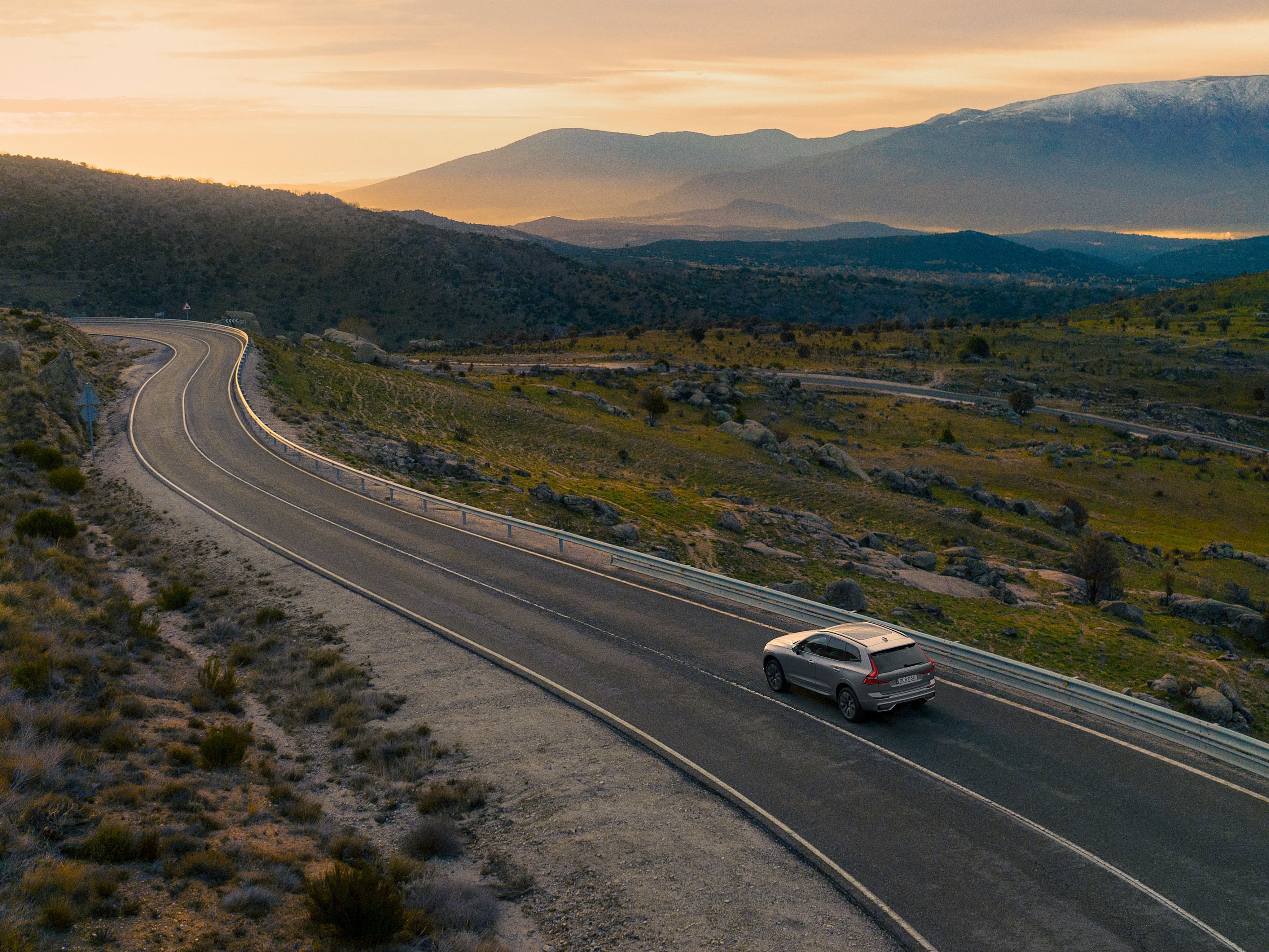 Een Volvo XC60 Recharge Plug-in hybrid rijdt op een bochtige weg bij zonsondergang.