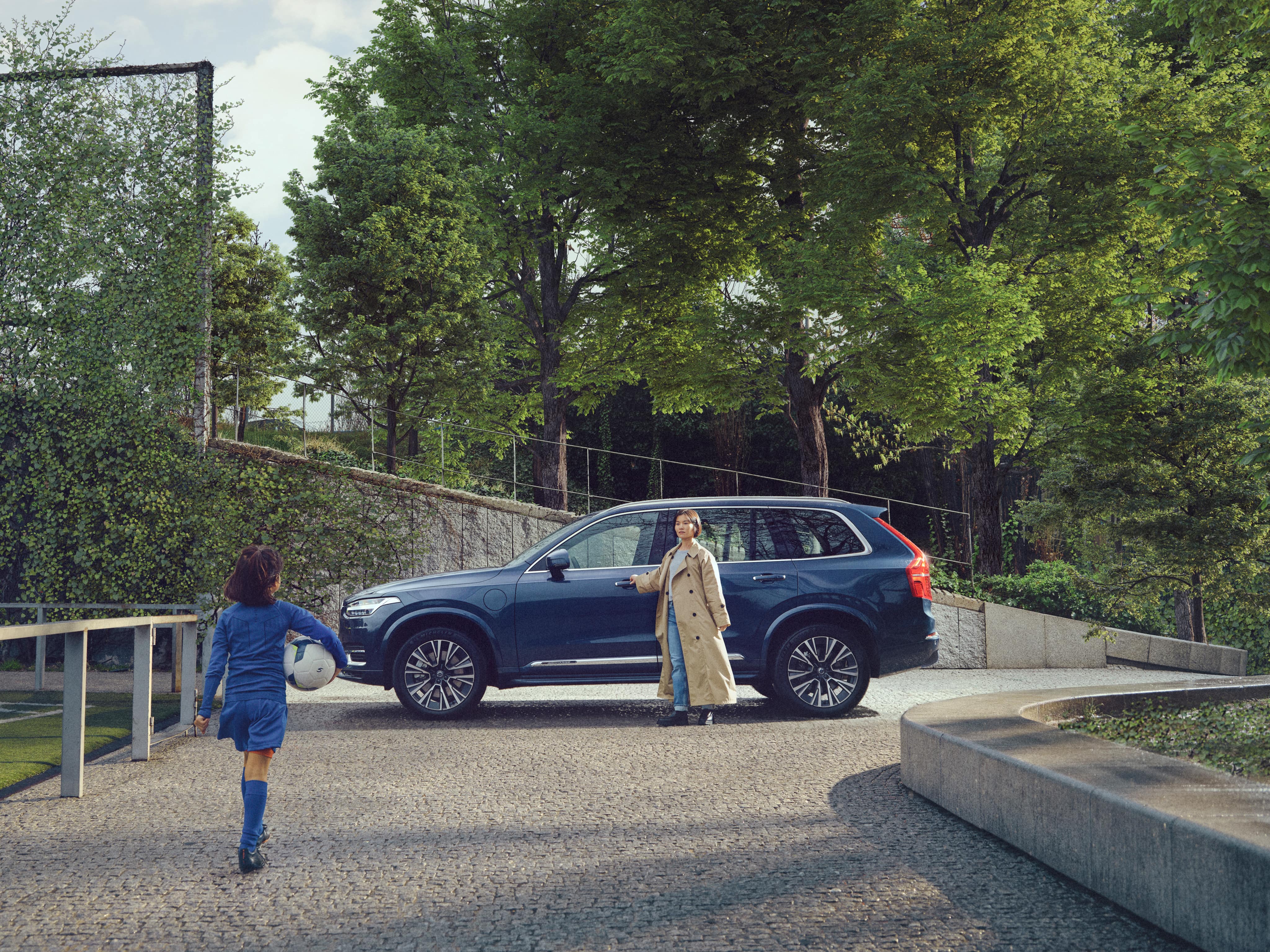 Young girl with a football walking towards her mother standing in front of a Volvo XC90 Recharge plug-in hybrid.