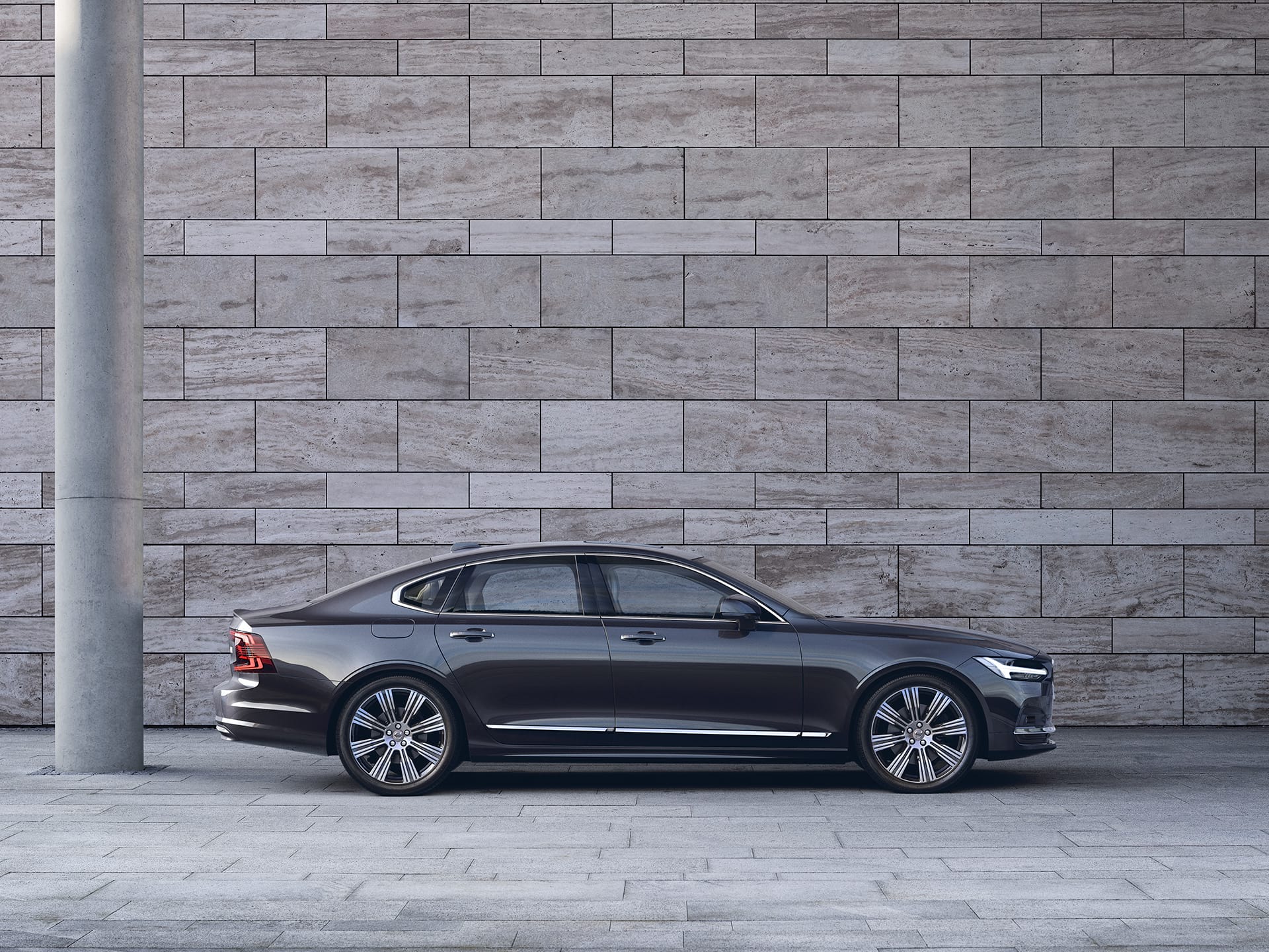 A dark Volvo S90 is parked in front of a grey wall.