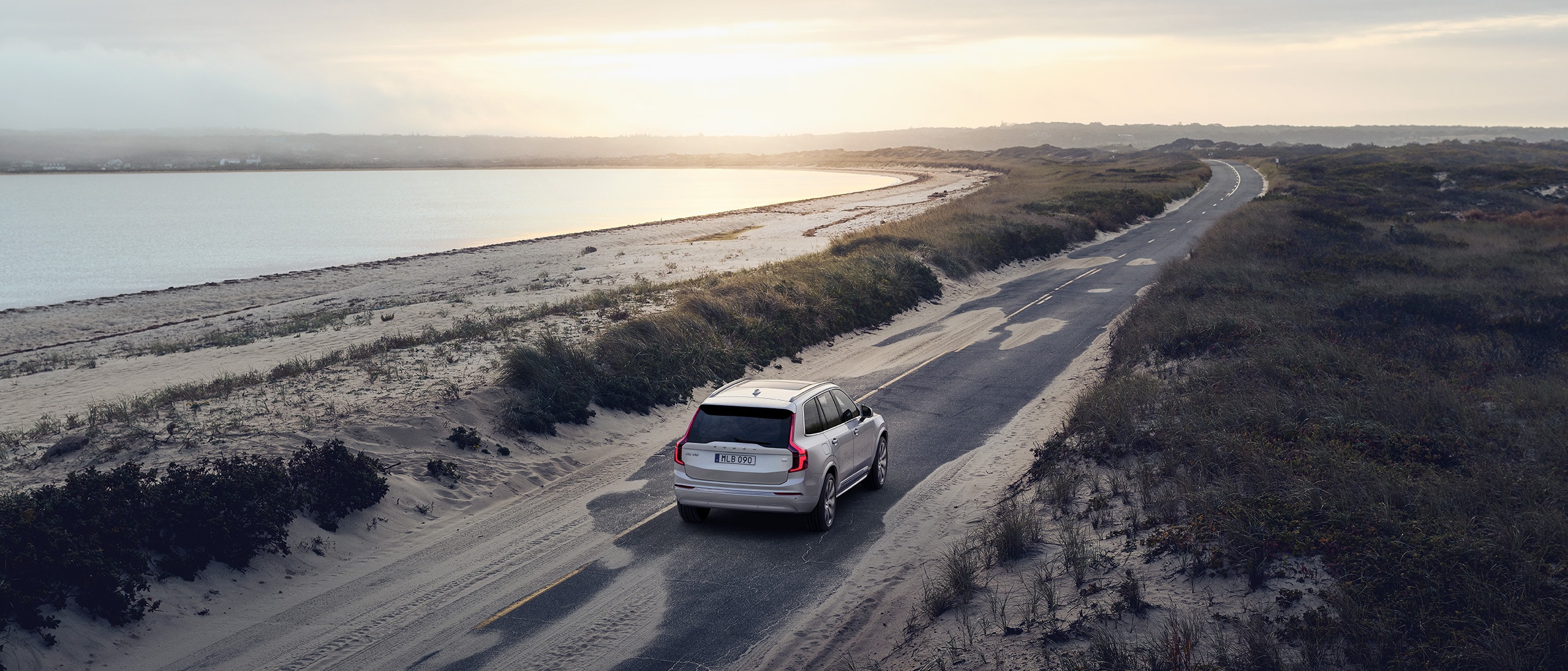 Ein grauer Volvo XC60 fährt in der Morgendämmerung auf einer Landstraße, die von Feldern umgeben ist 
