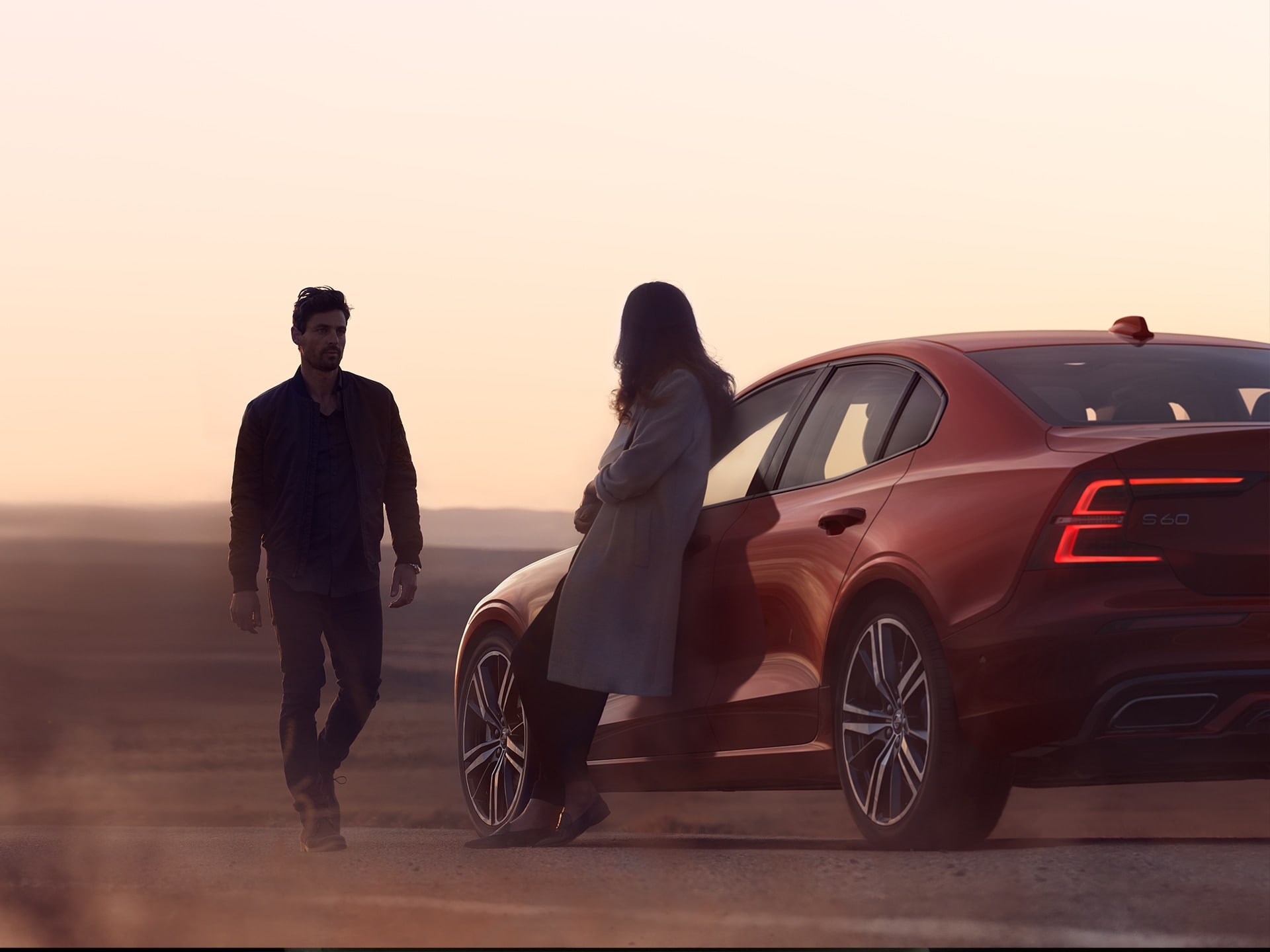 Un homme et une femme se tenant près de leur petite berline sportive Volvo S60 rouge au coucher du soleil.