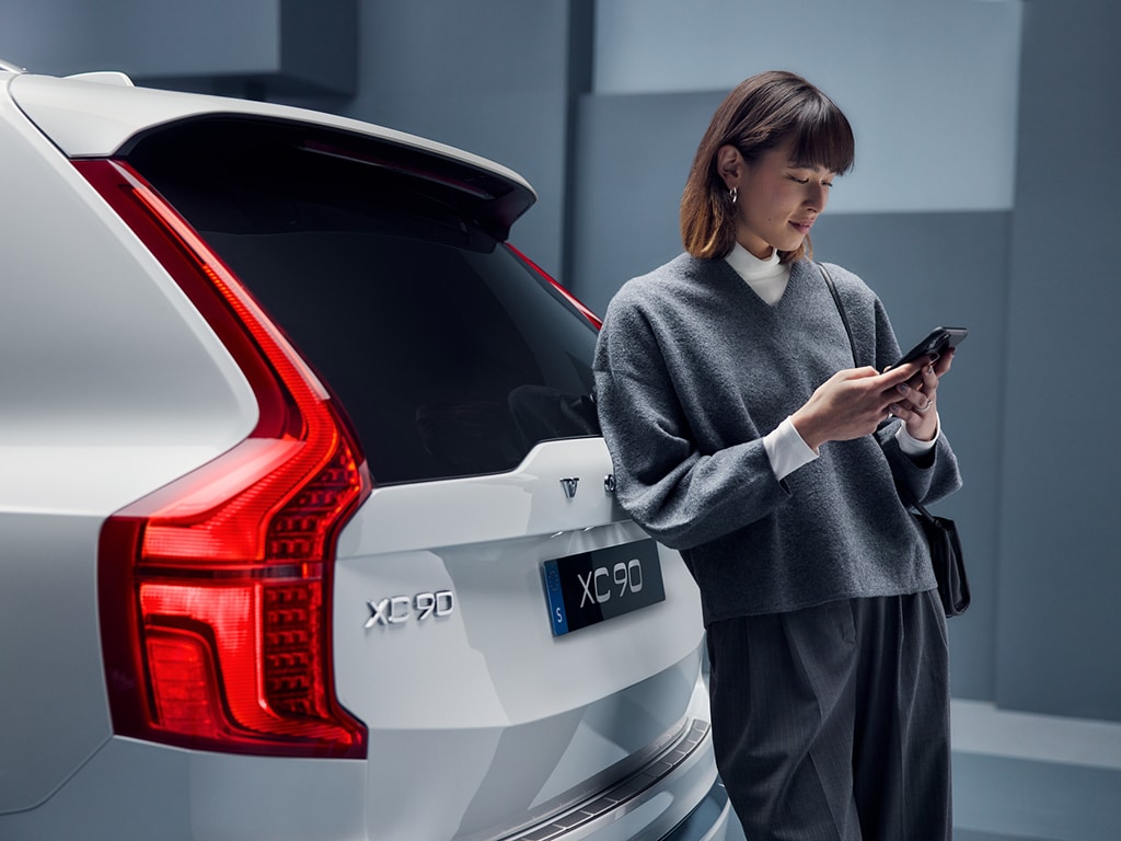 O doamnă stând alături de un Volvo XC90 argintiu și uitându-se la telefon.