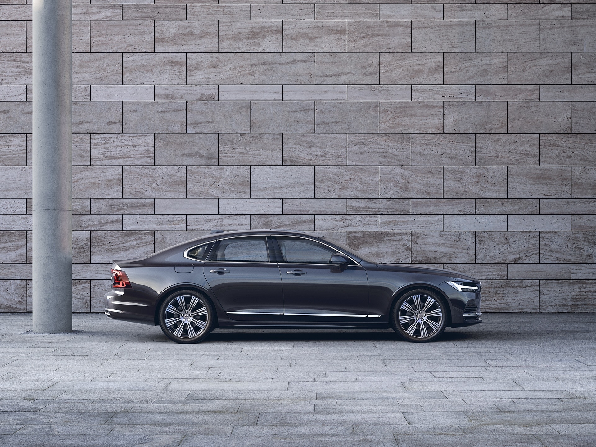 A dark Volvo S90 is parked in front of a grey wall.