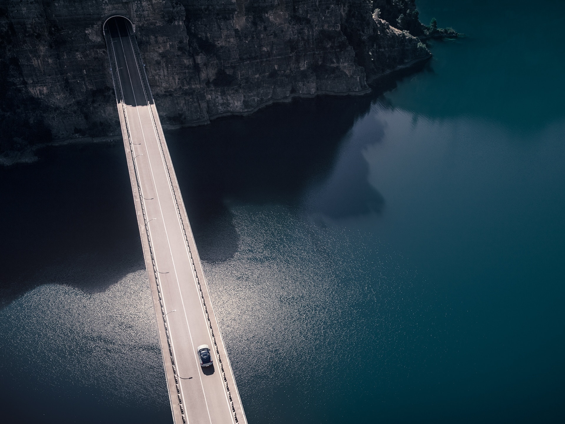 A Volvo XC40 Recharge drives on a bridge over the water that leads into a mountain tunnel.