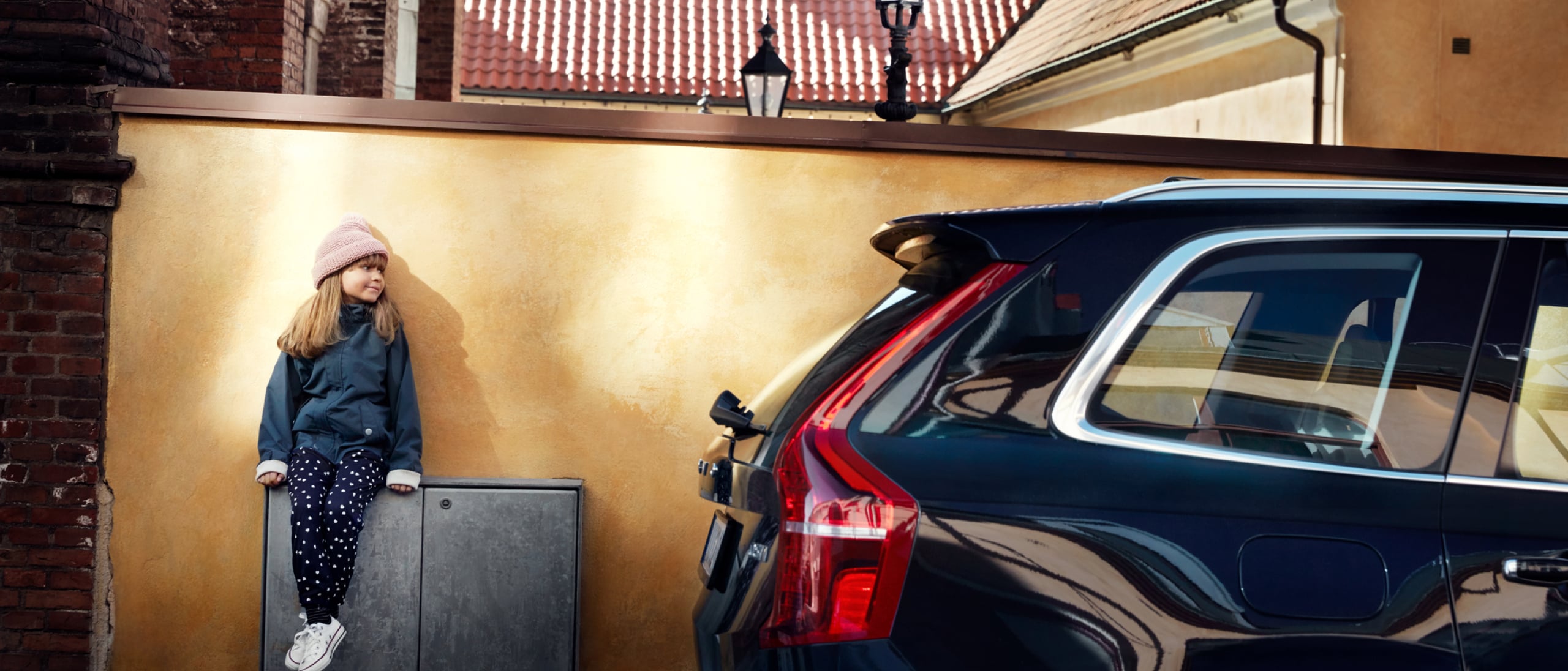 Et barn sidder lænet op ad en væg med en sort Volvo parkeret ved siden af.