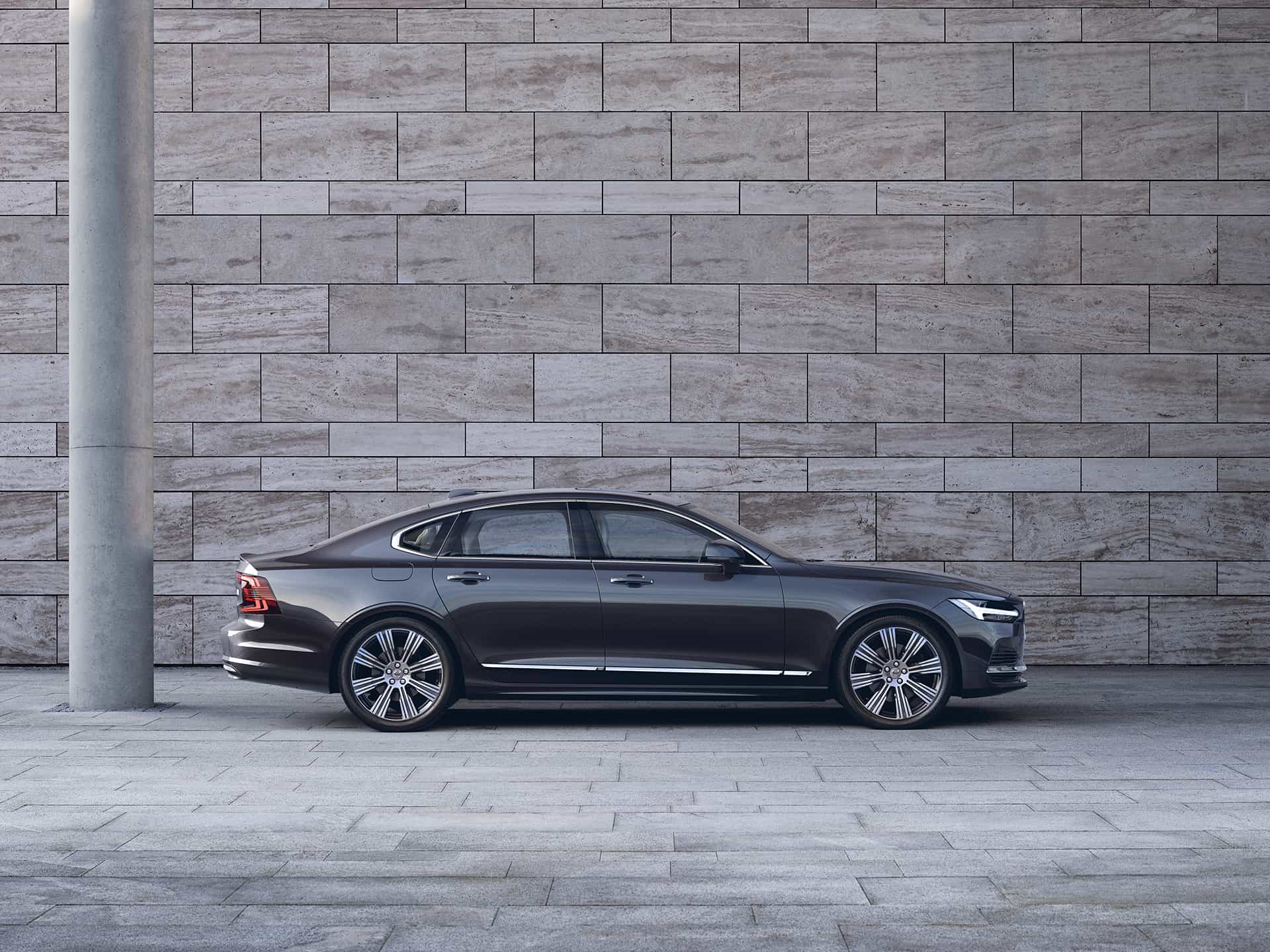A dark Volvo S90 sedan is parked in front of a grey wall.