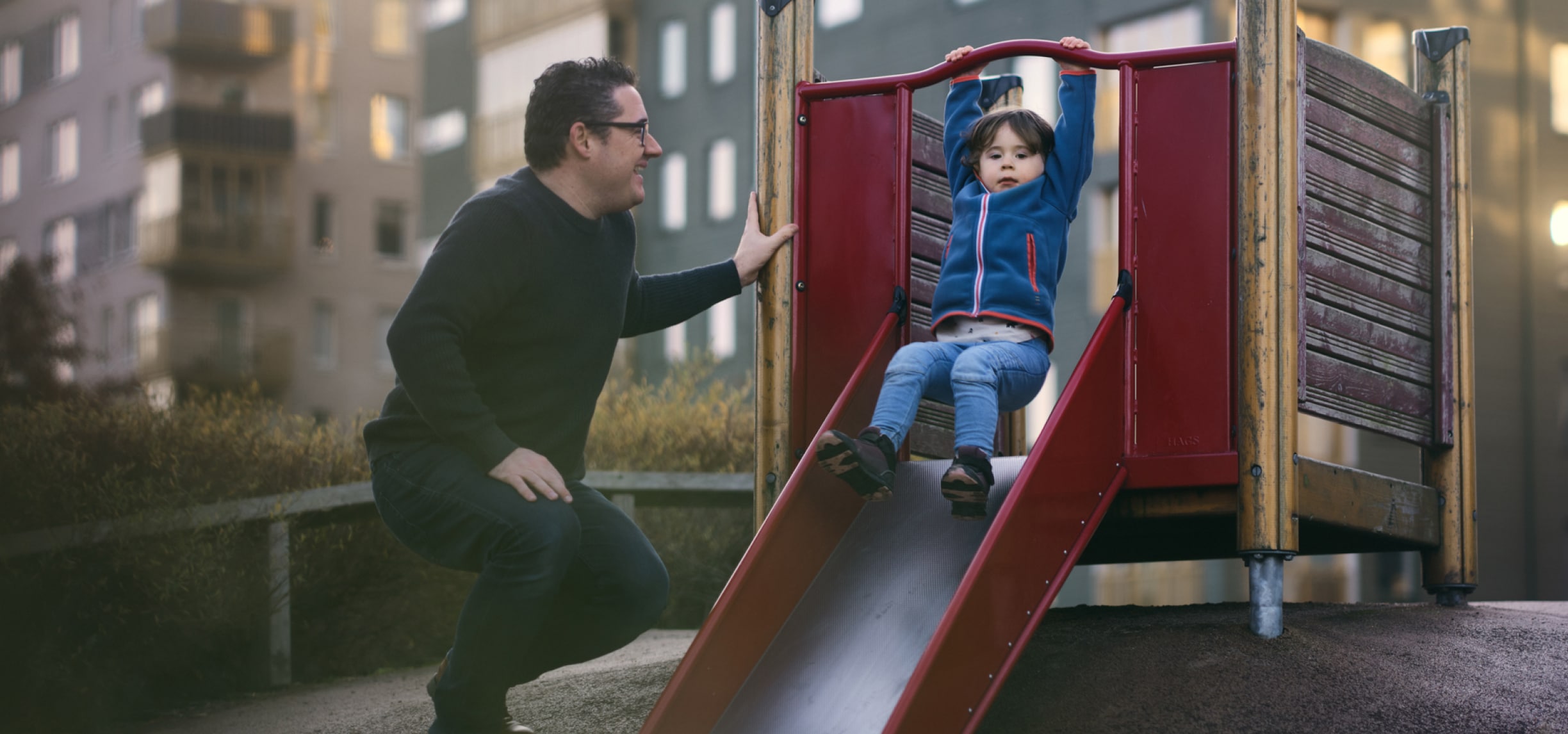 Um homem sorrindo para uma criança que brinca em um escorregador dentro de uma área de recreação infantil.