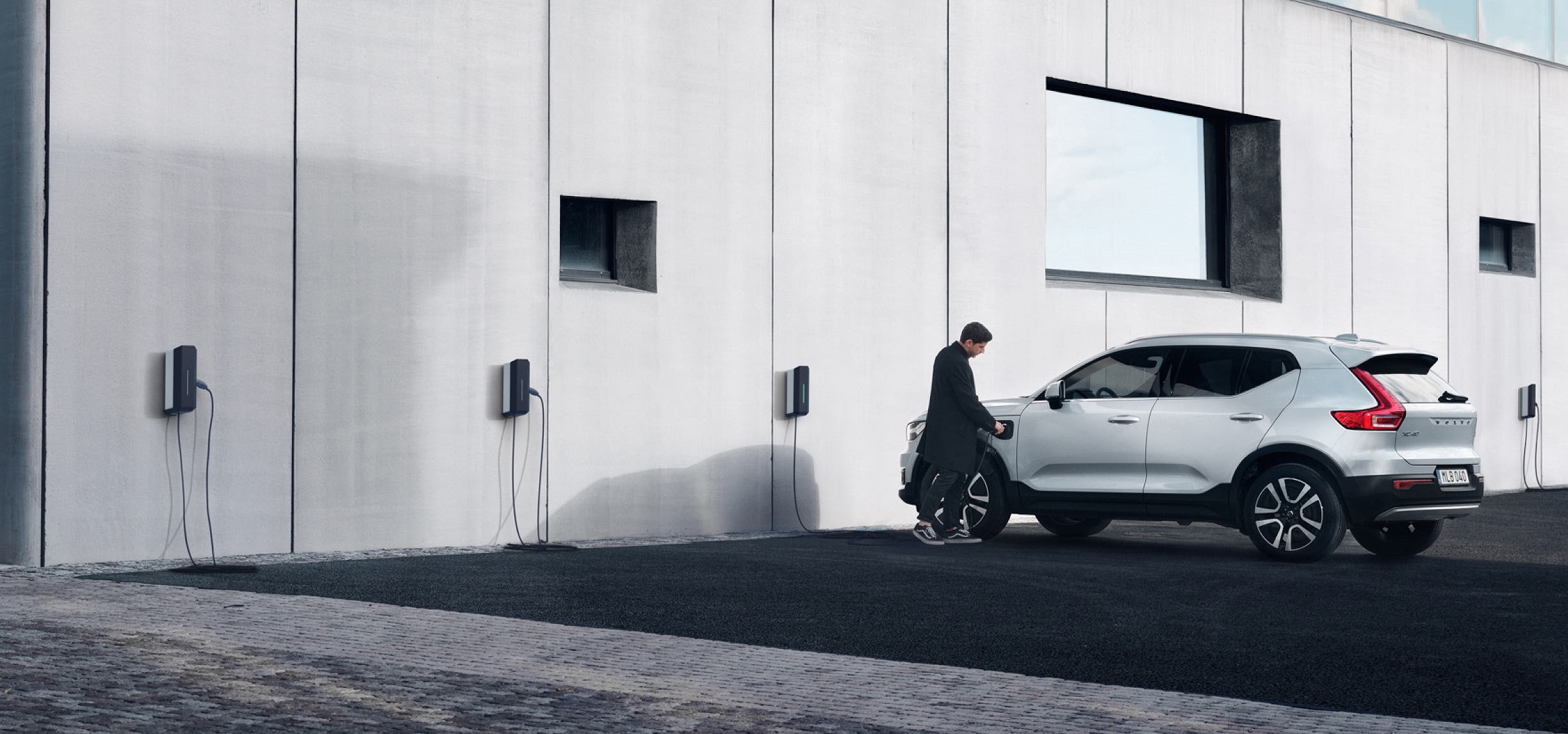 Um homem a ligar um carregador de parede a um Volvo XC40 cinzento estacionado junto a uma estação de carregamento