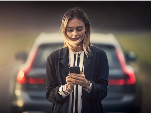 Woman standing in front of a new Volvo SUV, using a smartphone to access the Volvo Valet app