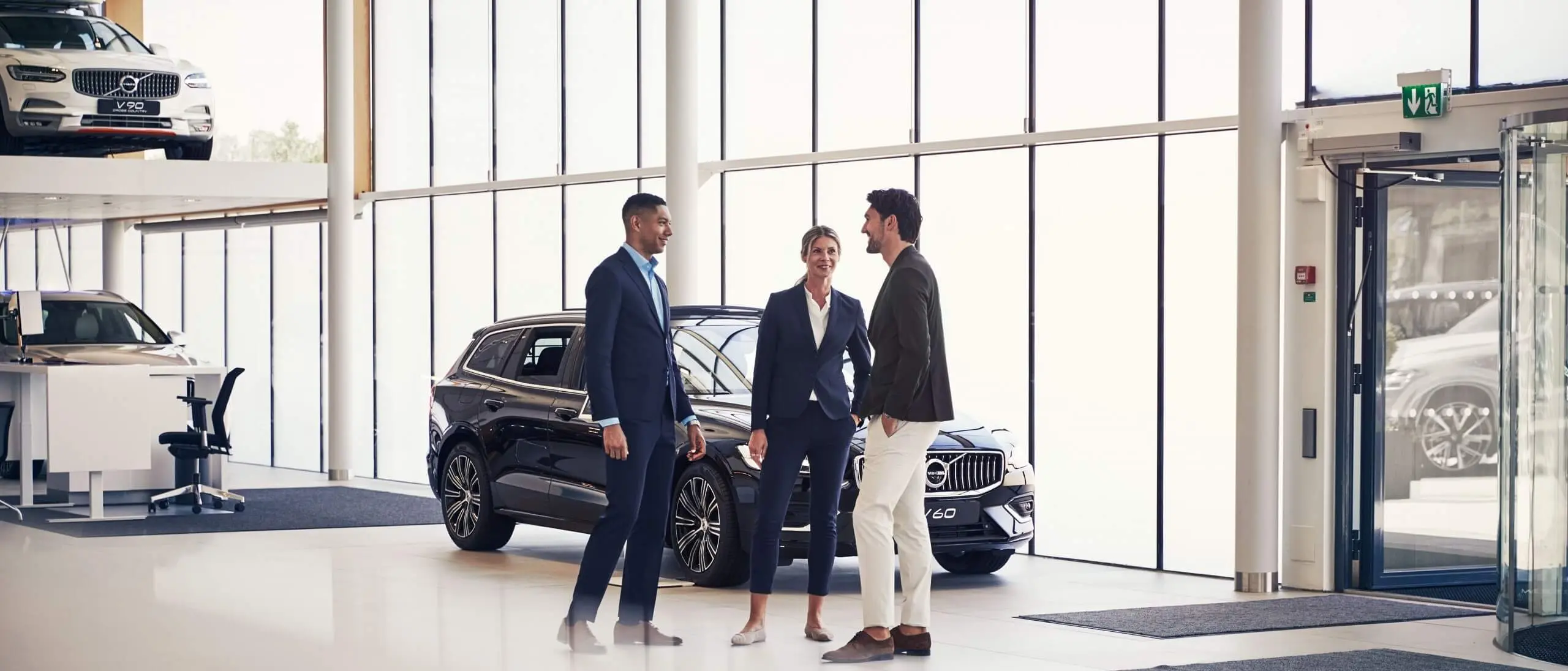 A Volvo dealer/retailer showroom in the background and a sales agent with custormers chatting.